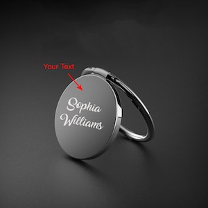 May include: A silver phone ring holder with the name "Sophia Williams" in white script. The ring is attached to a circular, gray disc with a swivel hinge and a silver ring for holding. The background is dark gray.