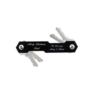 May include: Black leather key organizer with a personalized message that reads "Merry Christmas Dad" on one side and "We Love you Merry & Robert" on the other side. The organizer has a silver metal key ring and is holding a set of keys.