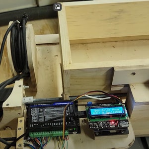 May include: A close-up of a woodworking machine with a blue LCD screen displaying "Box Joint Maker" and "Adj 1.112500". The machine has a black motor, a wooden frame, and a black cable spool.