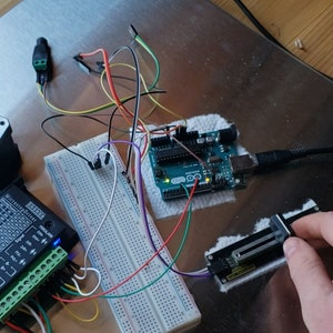 Può includere: Una scheda microcontrollore Arduino blu collegata a una breadboard e a un driver per motore passo-passo. Il driver è collegato a un motore passo-passo con una barra nera collegata ad esso. Un cavo nero collega l'Arduino a un computer.