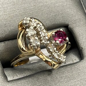 May include: A gold ring with a heart-shaped design featuring a red gemstone and small clear gemstones.