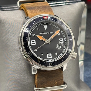 May include: A Morphic watch with a black face, white numerals, and orange hands. The watch has a stainless steel case and a brown leather strap. The watch is displayed on a gray pillow.