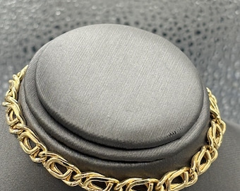 May include: A gold-colored chain bracelet with a repeating oval link design. The bracelet is displayed on a gray surface, showcasing its intricate pattern and polished finish. The jewelry piece is a classic accessory.