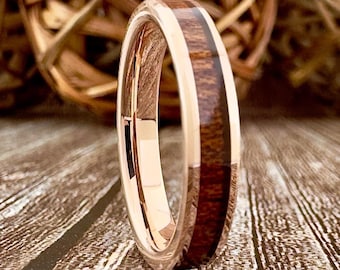 May include: Rose gold wedding band with a dark wood inlay. The ring features a polished finish and a contrasting wood grain pattern. The band is set against a wooden surface, highlighting the ring's design.
