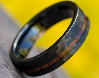 Puede incluir: Primer plano de un anillo negro con un acabado de metal martillado y una fina incrustación de madera marrón rojizo. El interior del anillo tiene un toque de azul y verde. El fondo es amarillo brillante.
