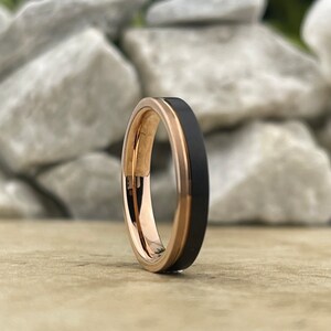 Puede incluir: Un anillo de carburo de tungsteno negro y oro rosa. El anillo tiene un borde biselado y un acabado pulido.