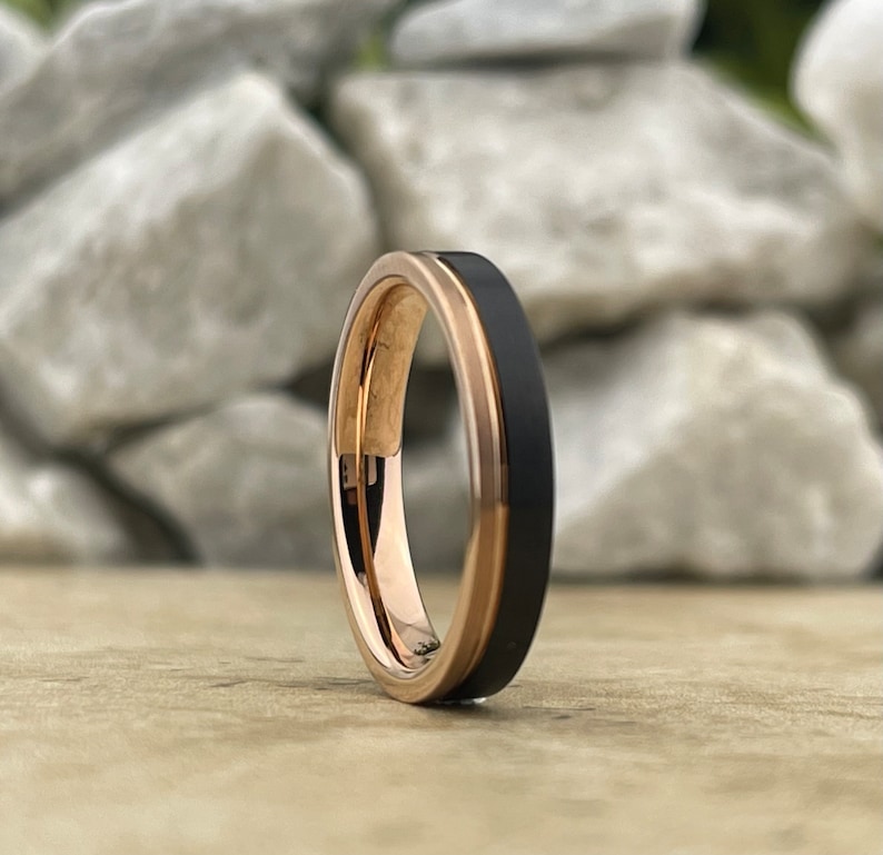 Puede incluir: Un anillo de carburo de tungsteno negro y oro rosa. El anillo tiene un borde biselado y un acabado pulido.