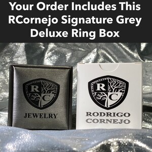 Puede incluir: Dos joyeros, uno gris y otro blanco, con el texto "RCornejo Signature Grey Deluxe Ring Box". La caja gris tiene la palabra "JEWELRY" y un logotipo. La caja blanca tiene el nombre "RODRIGO CORNEJO" y un logotipo.