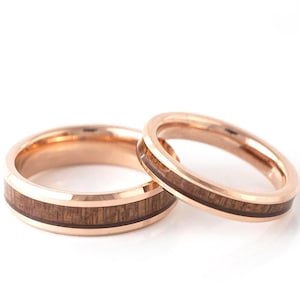 Puede incluir: Dos anillos de boda de oro rosa con incrustaciones de madera. Los anillos est&aacute;n sobre un fondo blanco. El anillo m&aacute;s grande tiene una banda m&aacute;s ancha que el anillo m&aacute;s peque&ntilde;o. La incrustaci&oacute;n de madera es de color marr&oacute;n oscuro.