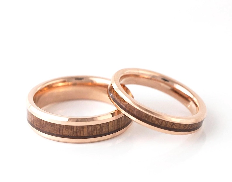 Puede incluir: Dos anillos de boda de oro rosa con incrustaciones de madera. Los anillos est&aacute;n sobre un fondo blanco. El anillo m&aacute;s grande tiene una banda m&aacute;s ancha que el anillo m&aacute;s peque&ntilde;o. La incrustaci&oacute;n de madera es de color marr&oacute;n oscuro.