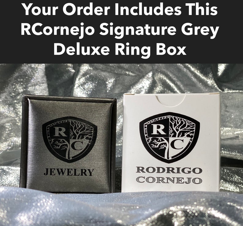 Puede incluir: Una caja de anillo gris con un logotipo en blanco y negro que dice "RCornejo Joyer&iacute;a". La caja est&aacute; cerrada y tiene una etiqueta blanca en el lateral que dice "Rodrigo Cornejo".
