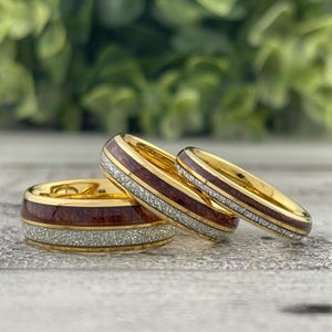 May include: Three gold rings with a red and white wood inlay. The rings are stacked on top of each other.