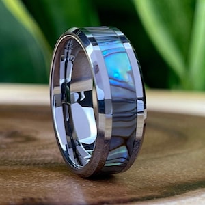 May include: A silver tungsten carbide ring with a blue and white abalone shell inlay. The ring has a bevelled edge and a polished finish.