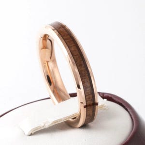 Puede incluir: Anillo de oro rosa con incrustaciones de madera marr&oacute;n oscuro. El anillo se encuentra sobre un fondo blanco, mostrando el metal pulido y la veta natural de la madera. Un primer plano resalta el dise&ntilde;o del anillo.