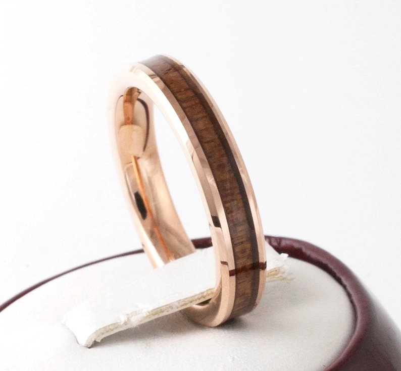 Puede incluir: Anillo de oro rosa con incrustaciones de madera marr&oacute;n oscuro. El anillo se encuentra sobre un fondo blanco, mostrando el metal pulido y la veta natural de la madera. Un primer plano resalta el dise&ntilde;o del anillo.