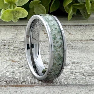 May include: A silver tungsten carbide ring with a green and white inlay. The ring is polished and has a beveled edge.