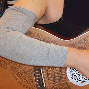 Peut inclure: Guitare acoustique avec une finition marron clair et un motif floral et de plumes de paon. La guitare est jouée, avec une manche grise couvrant le bras. La rosace présente un motif décoratif.