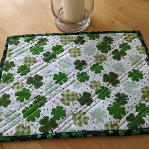 May include: A green shamrock patterned quilted placemat with a black border. The placemat is made of fabric and has a white background with green shamrocks and white polka dots.
