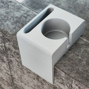 May include: A light grey couch armrest organiser featuring a circular cup holder and a rectangular slot for a remote control or smartphone. The organiser has a smooth, matte finish and is designed to drape over the side of a sofa armrest.