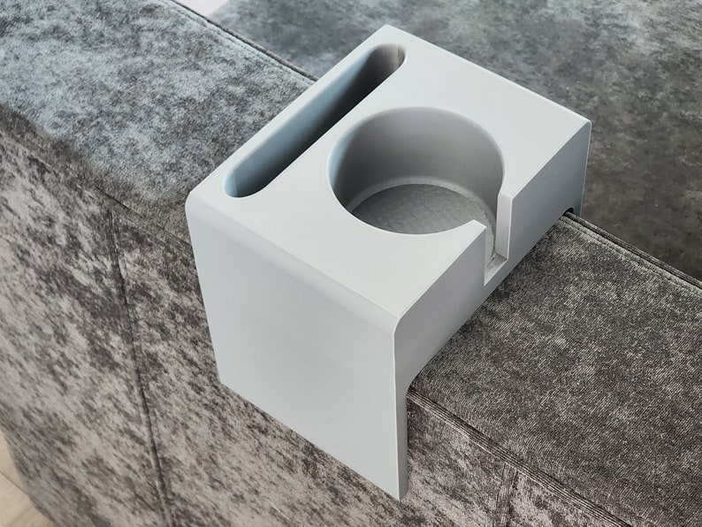May include: A light grey couch armrest organiser featuring a circular cup holder and a rectangular slot for a remote control or smartphone. The organiser has a smooth, matte finish and is designed to drape over the side of a sofa armrest.