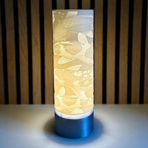 May include: A white ceramic table lamp with a cylindrical shade featuring an embossed design of koi fish. The lamp has a silver metal base.