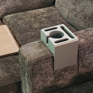 May include: A light grey, rectangular sofa armrest organiser featuring a central circular cup holder and three additional elongated slots for storage. The organiser is designed to drape over the arm of a couch for convenient access to items.
