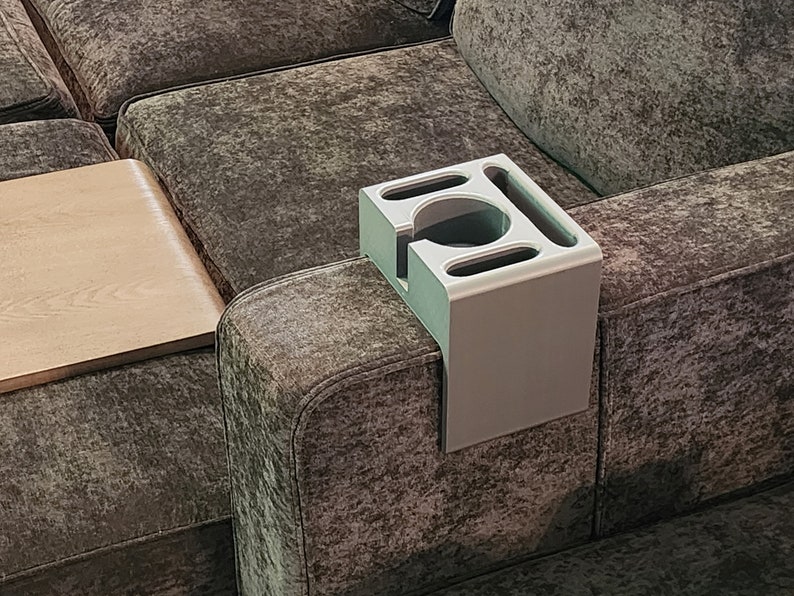 May include: A light grey, rectangular sofa armrest organiser featuring a central circular cup holder and three additional elongated slots for storage. The organiser is designed to drape over the arm of a couch for convenient access to items.