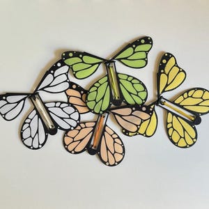 May include: Five colorful paper butterflies with black outlines and a stick in the middle. The butterflies are in shades of green, yellow, orange, and white.