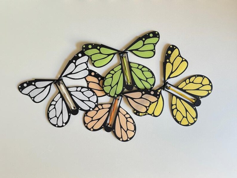 May include: Five colorful paper butterflies with black outlines and a stick in the middle. The butterflies are in shades of green, yellow, orange, and white.