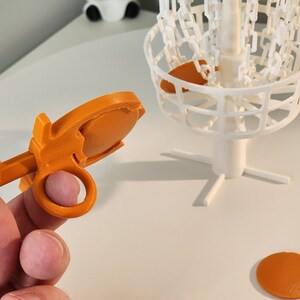 Puede incluir: Objetivo de disco de golf de pl&aacute;stico naranja con una cesta de metal blanca y una base blanca. El objetivo se sostiene en una mano.