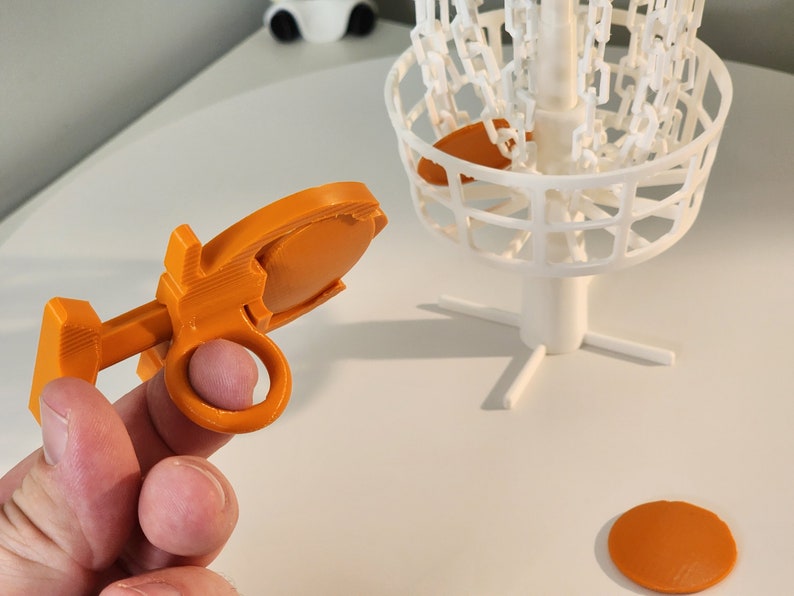 Puede incluir: Objetivo de disco de golf de pl&aacute;stico naranja con una cesta de metal blanca y una base blanca. El objetivo se sostiene en una mano.