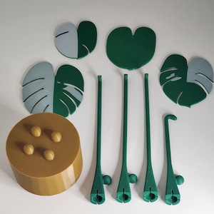 May include: A set of five green and gray leaf-shaped plant stakes with a brown base with four round knobs. The stakes are designed to look like monstera leaves.