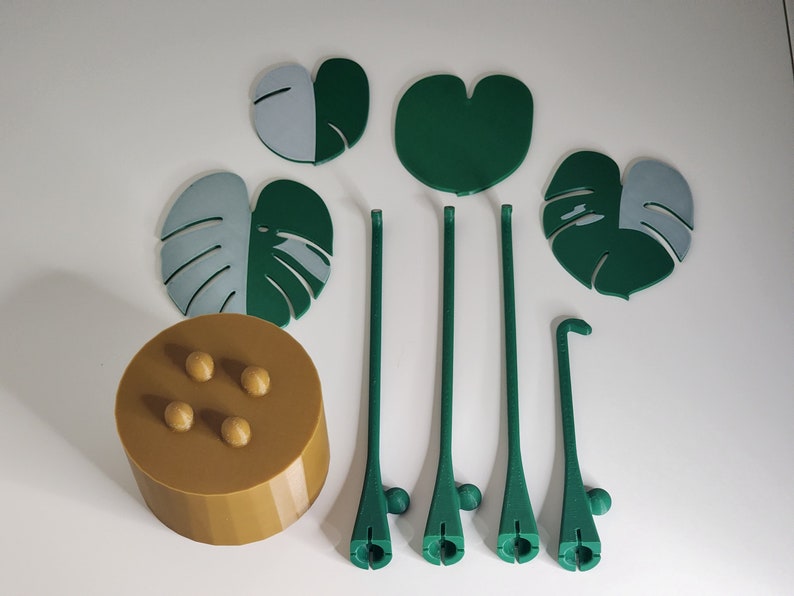 May include: A set of five green and gray leaf-shaped plant stakes with a brown base with four round knobs. The stakes are designed to look like monstera leaves.