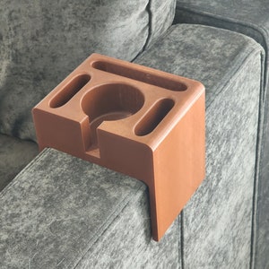 May include: A terracotta-coloured sofa armrest organiser featuring a central circular cup holder, a long rectangular slot, and two smaller oval-shaped compartments. The organiser has a rigid, L-shaped design that fits over the arm of a sofa.