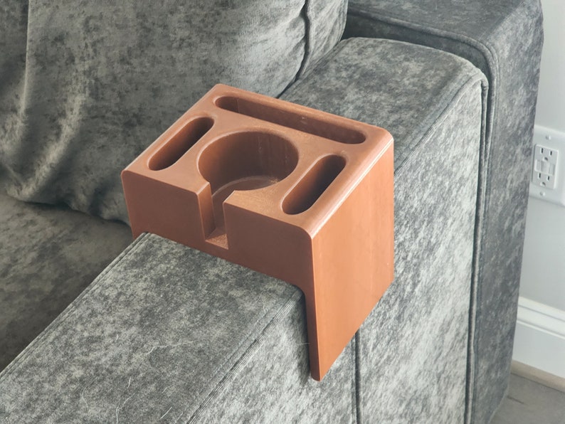 May include: A terracotta-coloured sofa armrest organiser featuring a central circular cup holder, a long rectangular slot, and two smaller oval-shaped compartments. The organiser has a rigid, L-shaped design that fits over the arm of a sofa.