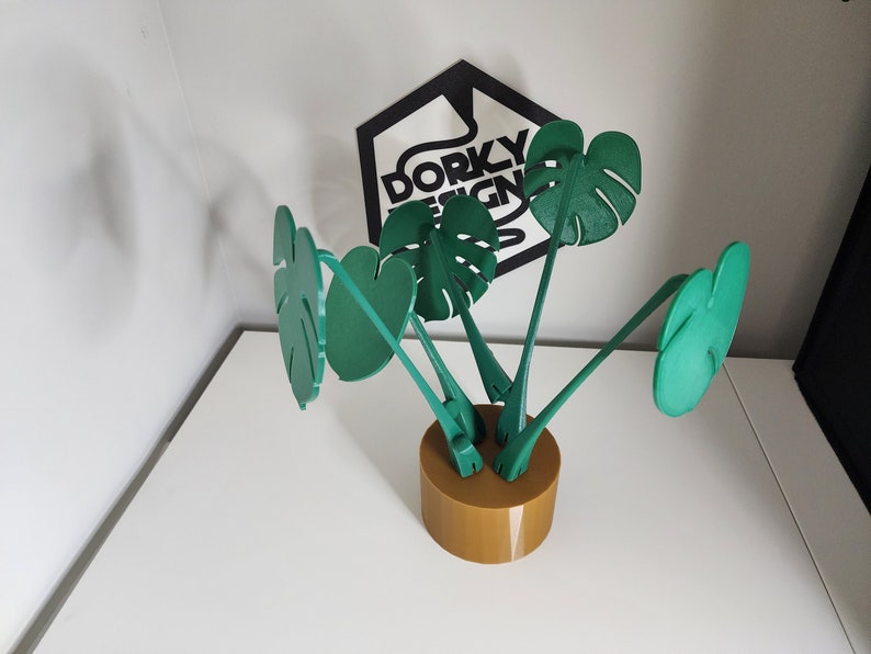 May include: A green, 3D printed, tabletop plant sculpture with four leaves shaped like monstera plants. The sculpture is mounted on a brown, cylindrical base.