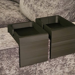May include: Two black, rectangular, L-shaped cup holders designed to drape over the arm of a sofa. Each holder features a flat, square-shaped recessed area for a drink or small item, with a smooth, matte finish and visible horizontal layer lines.