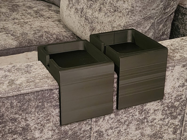 May include: Two black, rectangular, L-shaped cup holders designed to drape over the arm of a sofa. Each holder features a flat, square-shaped recessed area for a drink or small item, with a smooth, matte finish and visible horizontal layer lines.