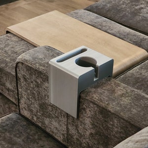 May include: A light grey, L-shaped sofa armrest organiser made of a smooth, matte material. It features a circular cutout for a cup, a rectangular slot for a mobile device, and a vertical notch on the front face for cable management.