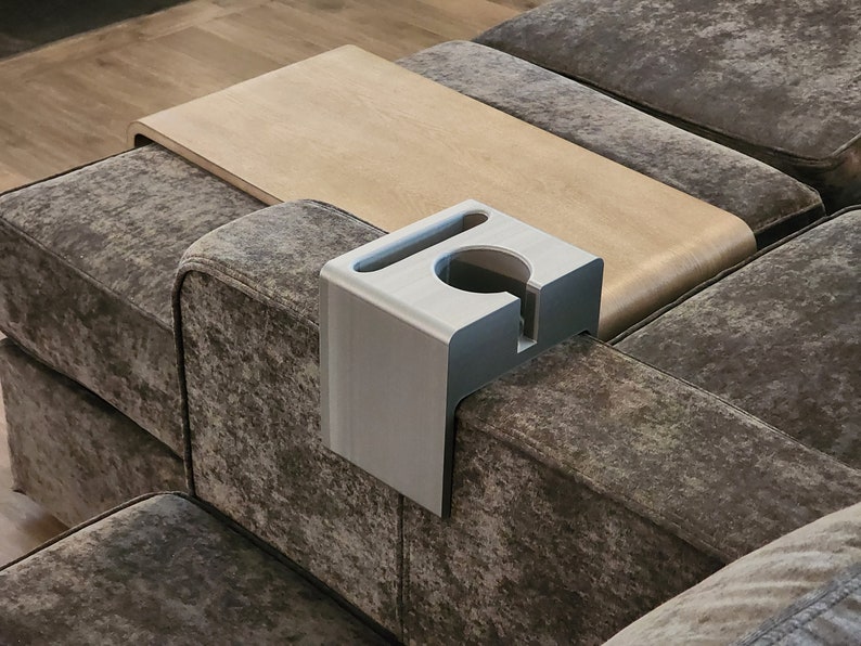 May include: A light grey, L-shaped sofa armrest organiser made of a smooth, matte material. It features a circular cutout for a cup, a rectangular slot for a mobile device, and a vertical notch on the front face for cable management.