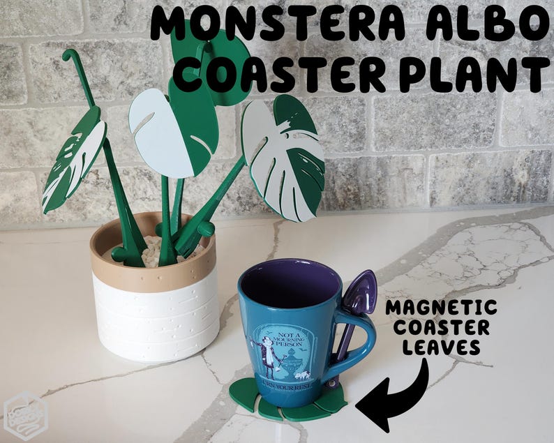 May include: A white ceramic planter with a brown rim holds a faux Monstera Albo plant with green and white leaves. The plant is displayed on a white countertop with the text "Monstera Albo Coaster Plant" and "Magnetic Coaster Leaves" visible.