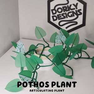Puede incluir: Una planta de pothos de papel verde y blanco con el texto "Dorky Designs" y "Pothos Plant Articulating Plant" sobre una superficie blanca.