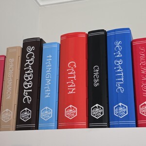 May include: A collection of board game boxes, including "Checkers," "Backgammon," "Scrabble," "Hangman," "Catan," "Chess," "Sea Battle," and "Four in a Row." Each box is a different color and has the game title printed on the spine.