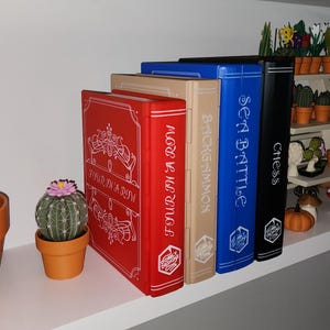 May include: A shelf displays decorative board game boxes labeled "Four in a Row," "Backgammon," "Sea Battle," and "Chess." A cactus and a small potted cactus are on the left. Miniature pumpkins and a white pitcher are on the right.