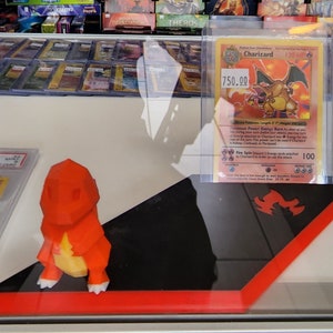 May include: A display case featuring a Charizard Pokémon card with a price tag of $750.00. An orange 3D-printed Charizard figure is on a black and red display stand. Other Pokémon cards are visible in the background.