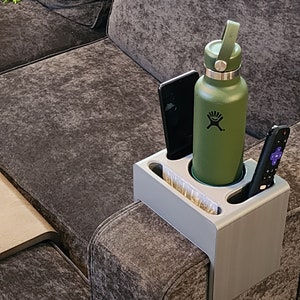 May include: A light grey sofa armrest organiser featuring a central circular cup holder containing a matte olive green Hydro Flask water bottle, a rectangular slot holding a black smartphone, and a narrow slot containing a black television remote.