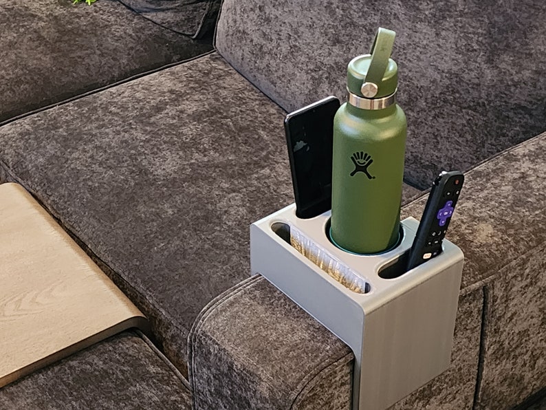 May include: A light grey sofa armrest organiser featuring a central circular cup holder containing a matte olive green Hydro Flask water bottle, a rectangular slot holding a black smartphone, and a narrow slot containing a black television remote.