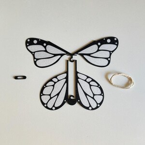 May include: Black paper butterfly craft with white wings and a black body. The butterfly is in pieces, with the wings separate from the body. There is a small black loop and a piece of string included.
