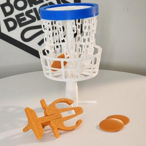 Puede incluir: Una cesta de disco golf de pl&aacute;stico blanco con un borde azul y una red de cadena. Hay tres discos de pl&aacute;stico naranja sobre una superficie blanca.