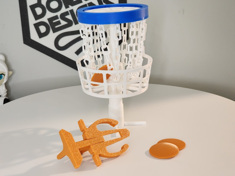 Puede incluir: Una cesta de disco golf de pl&aacute;stico blanco con un borde azul y una red de cadena. Hay tres discos de pl&aacute;stico naranja sobre una superficie blanca.